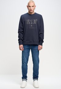 Jūras spēku kreklis ar lielu pelēku "BIG STAR" tekstu un saukli, kopā ar zilu džinsu un baltām sporta apaviem. Ikdienišķs, brīvs piegriezums un apaļa kakla izgriezums.