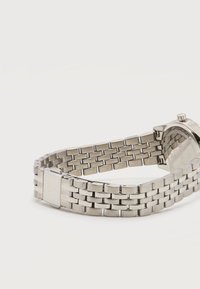 Ρολόι από ανοξείδωτο ατσάλι με μεταλλικό μπρασελέ με συνδέσμους και αγκράφα χαραγμένη με τη λέξη «BOSS», εμφανίζεται από πίσω σε λευκή επιφάνεια.