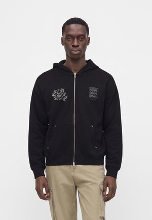 Junger Mann, der einen schwarzen Zip-up-Hoodie mit grauen Rosen- und Schildemblemen und beige Cargohosen trägt, steht vor einem einfachen weißen Hintergrund.