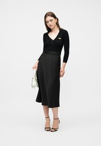 Γυναίκα που φοράει μαύρο μακρυμάνικο τοπ και μαύρη midi φούστα, κρατώντας μια λευκή τσάντα χειρός, στέκεται με μαύρες πέδιλα με λουράκια σε απλό φόντο.