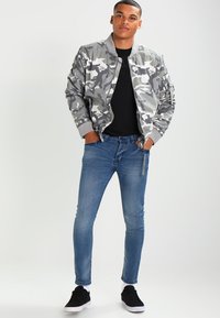 Chaqueta bomber de camuflaje gris con puños acanalados, combinada con jeans ajustados azul y zapatillas negras. Diseño simple y funcional.