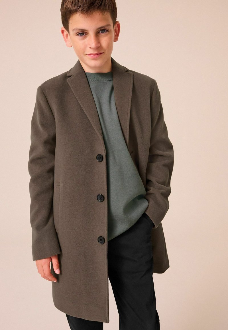 Lungo cappotto verde oliva realizzato in tessuto morbido, caratterizzato da revers a punta, tre bottoni neri e una vestibilità comoda sopra un top grigio e pantaloni neri.
