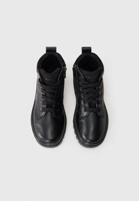 Une paire de bottes noires en cuir à lacets avec des fermetures éclair latérales et des boucles, vue du dessus sur un fond clair.