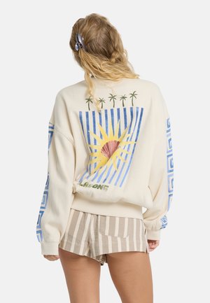 Donna con capelli biondi che indossa una felpa color crema con grafica di palma e sole, abbinata a pantaloncini beige e bianchi a righe, in piedi con la schiena rivolta alla fotocamera.