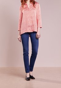Rød og hvid mønstret langærmet bluse med V-hals, iført mørkeblå skinny jeans og sorte sko, mod en ensfarvet baggrund.