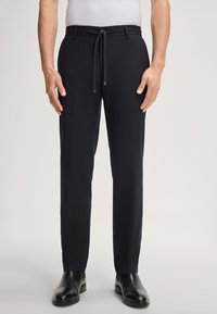 Man in een zwart slim-fit broek met een trekkoord in de taille en zwarte leren schoenen tegen een effen achtergrond.