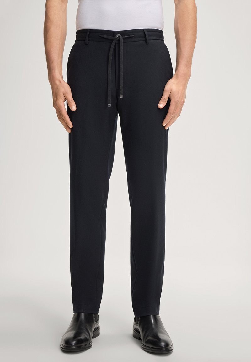 Man in een zwart slim-fit broek met een trekkoord in de taille en zwarte leren schoenen tegen een effen achtergrond.