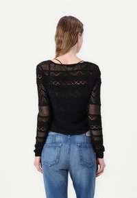 Czarna, siateczkowa bluzka z długim rękawem z geometrycznymi wzorami i przezroczystą fakturą, zestawiona ze średniodniowymi niebieskimi jeansami z tylnymi kieszeniami.