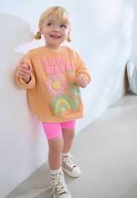 Sudadera color durazno con texto "SURF WAVES", gráficos en rosa y verde, combinada con shorts cortos de canalé rosa. Zapatillas altas blancas con acentos de purpurina.