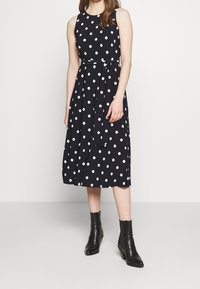 Černé bezrukávové šaty s bílými puntíky, ke kolenům, stažené v pase páskem. Modelka má na sobě černé kotníkové boty.