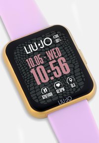 Reloj inteligente cuadrado con una pantalla negra que presenta un patrón texturizado, acentos dorados y una correa de silicona rosa. Muestra la hora y métricas de salud.