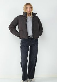 Schwarze gepolsterte Jacke mit hohem Kragen, mit einem Reißverschluss vorne, seitlichen Taschen und einem lockeren Schnitt, kombiniert mit dunklen Jeans und Sneakers.