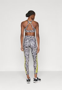 Σετ αθλητικό σουτιέν και κολάν σε γκρι χρώμα με λευκά μοτίβα κειμένου. Η πλάτη διαθέτει σχεδιασμό με άνοιγμα. Το κολάν έχει κίτρινες πλευρικές ρίγες και εφαρμοστή γραμμή.