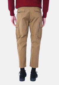 Pantaloni cargo in cotone beige con due tasche laterali, due tasche posteriori, una vestibilità rilassata e orli arrotolati. Indossati con scarpe nere e calze.