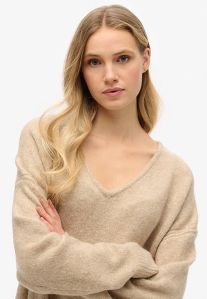 Maglione beige a maglia con scollatura a V e maniche ampie. Il materiale sembra morbido con una finitura fine. Il modello ha capelli biondi mossi e le braccia incrociate.
