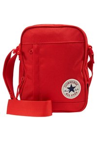 Červená kompaktní taška přes rameno s nastavitelným popruhem, přední kapsou na zip a kulatou bílou nášivkou s logem Converse All Star.