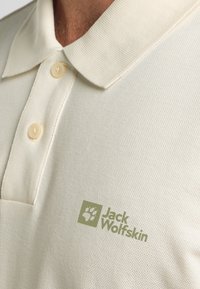  Lichtbeige poloshirt van een gestructureerde stof, met een klassieke kraag, twee knopen en een groen logo aan de linkerkant.