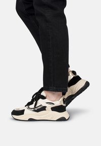Sneakers beige e neri con suola spessa, caratterizzati da un mix di pelle e tessuto, lacci neri e un design texturizzato.