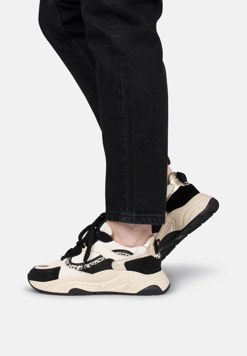 Sneakers beige e neri con suola spessa, caratterizzati da un mix di pelle e tessuto, lacci neri e un design texturizzato.