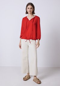 Blusa leggera rossa a maniche lunghe con scollatura a V e orlo con coulisse, abbinata a pantaloni bianchi larghi e scarpe slip-on color beige con dettagli a fiocco.
