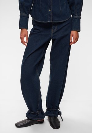 Personne portant un pantalon en denim bleu foncé avec des ourlets à cordon de serrage et des chaussures noires sans lacets, assortie d'une veste en jean assortie.