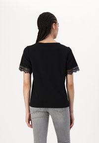 Svart T-skjorte med korte ermer, med blondebeskrivelse på ermene, laget av et glatt stoff. Bakvisning viser en rund hals.