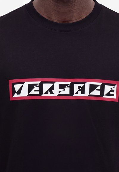 Chemise noire avec un texte géométrique stylisé en blanc et noir épelant "Versace" à l'intérieur d'une bordure rectangulaire rouge sur la poitrine.