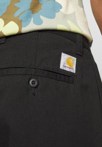 Zbliżenie na tylnią kieszeń czarnych spodni Carhartt z guzikiem i białą naszywką z logo, w połączeniu z wielokolorową koszulą w kwiaty.