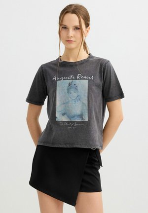 Mujer joven con cabello castaño claro en una coleta, vestida con una camiseta negra descolorida con gráfico de Auguste Renoir y una falda negra asimétrica.