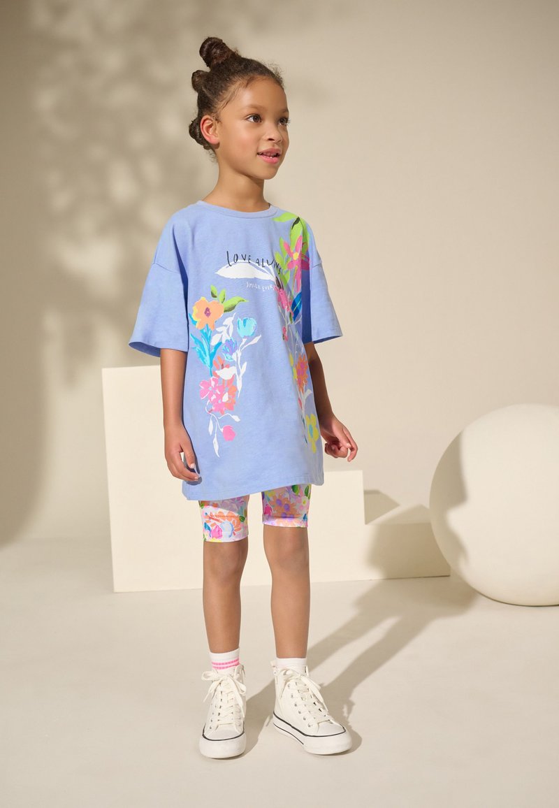 T-shirt bleu oversize avec des motifs floraux, associée à un short de vélo coloré à motifs et des baskets montantes blanches. Fond uni.