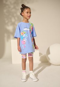 Kék, túlméretezett póló virágmintákkal, színes mintás biciklis rövidnadrággal és fehér magas szárú tornacipőkkel párosítva. Egyszínű háttér.