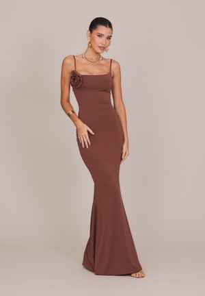 Femme portant une robe ajustée marron sans manches avec un détail floral sur la poitrine, des bijoux en or et des sandales, debout devant un fond uni.