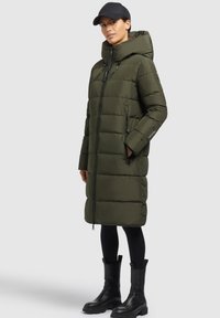 Mujer con un abrigo de plumas largo color verde oliva y capucha, leggings negros, botas altas negras y una gorra de béisbol negra, de pie contra un fondo blanco.
