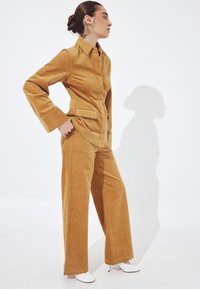 Ženska stoji bočno, oblečena v gorčično kordurojno jakno in hlače, obuta v bele pete, na belem ozadju, ki meče senco.