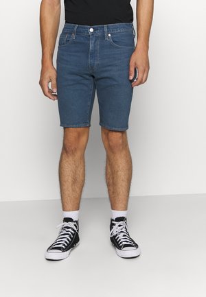 Shorts en denim, bleu foncé, longueur mi-cuisse, design classique à cinq poches avec une fermeture à bouton métallique, associés à des baskets montantes noires.