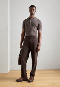 Polo shirt marron à manches courtes avec détails de boutons, associé à une pantalon marron foncé et des chaussures à lacets marron. Sac à bandoulière marron inclus.