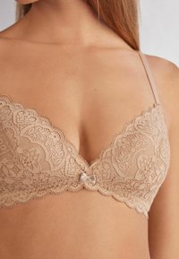 Beżowy stanik koronkowy z kwiatowym wzorem, głębokim dekoltem i delikatną kokardką na środku. Regulowane ramiączka i falbaniaste krawędzie.