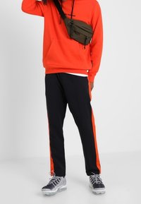 Sudadera roja con bolsillo frontal y pantalones de chándal negros con rayas laterales naranjas. Combinados con zapatillas grises y un bolso cruzado en color oliva.