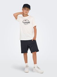 White t-shirt with a black graphic print, paired with dark shorts and white sneakers. Background is light gray. Boy poses casually.