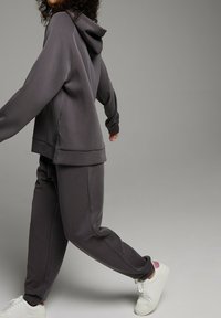 Szara bluza z kapturem i pasujące dresy wykonane z miękkiego materiału, z luźnym krojem i subtelnymi detalami po bokach, w połączeniu z białymi sneakersami.