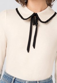 Kvinna med en krämfärgad långärmad topp med en rund krage kantad med svarta volanger som är knutna med ett svart band, tillsammans med blå denimjeans.