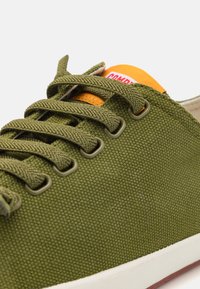 Grüne Leinen-Sneaker mit runder Zehenpartie, weißer Gummisohle, flachen Schnürsenkeln und orangefarbenem Etikett auf der Zunge. Besitzt ein strukturiertes Gewebe und Metallösen.