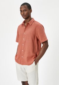 Camisa de lino coral de manga corta con botones en el frente, cuello extendido y un corte relajado, combinada con pantalones cortos de color claro.