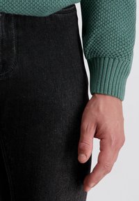 Grüner strukturierter Strickpullover mit gerippten Bündchen, kombiniert mit einer dunklen Jeans. Nahaufnahme einer Hand, die auf dem Hosenbein ruht und die Stoffdetails zeigt.