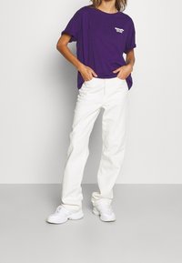 Lila bomull t-shirt med rullade ärmar och vit tryck på bröstet, tillsammans med vita denimbyxor och vita sneakers.