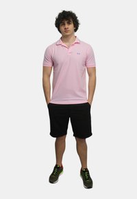Polo shirt rosa chiaro con maniche corte e colletto, che presenta un piccolo logo blu. Abbinato a pantaloni corti neri e sneakers verdi.
