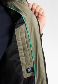 Futter einer gepolsterten Jacke aus olivgrünem und schwarzem Nylonstoff, mit türkisfarbenen Paspeln und einem Etikett mit dem Branding "Parode Industries".
