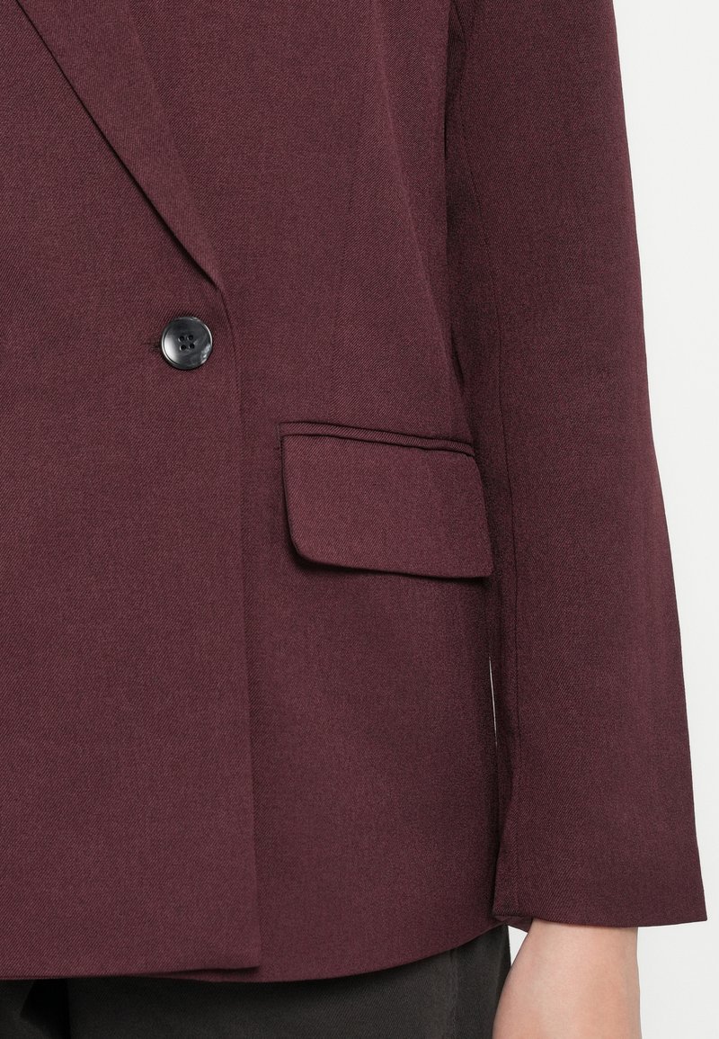 Blazer borgogna con rever a punta, bottone nero singolo e tasca laterale in un tessuto liscio e testurizzato.