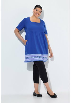 Smilende kvinde i blå kortærmet tunika med hvidt og lilla mønster ved kanten, sorte leggings og sorte balletsko, stående mod en hvid baggrund.