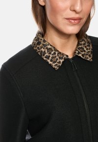 Black zip-up jacket with a textured fabric. Features a leopard print collar in shades of brown and black. Smooth finish and fitted design.